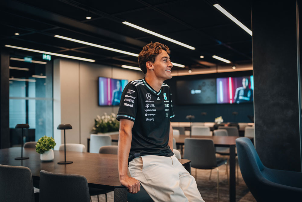 George Russell, Mercedes-AMG PETRONAS Formula One Team’in pilotu, ekibin modern ve şık misafir alanında veya ofisinde bir masanın üzerinde rahat bir şekilde oturuyor. Gülümsüyor ve üzerinde Einhell, IWC ve CrowdStrike gibi sponsor logolarının yer aldığı resmi takım gömleğini giyiyor. Arka plandaki ekranlarda onun görüntüsü yer alıyor.