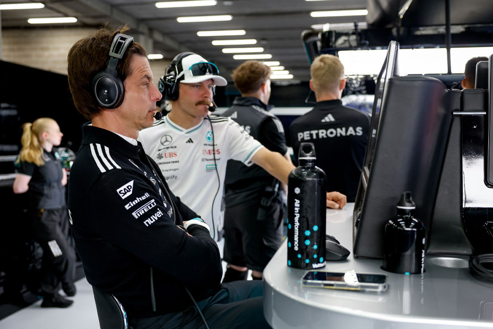 Mercedes-AMG PETRONAS Formula One Team’den Toto Wolff ve Valtteri Bottas, garajdaki ekip çalışma istasyonunda görülüyor. Yarış stratejisine odaklanmış durumdalar, ekranları izliyor ve kulaklıklarla iletişim kuruyorlar.