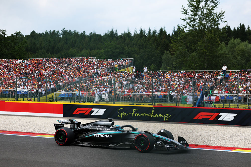 Mercedes-AMG PETRONAS Formula One Team’e ait bir araç Spa-Francorchamps pistinde yarışıyor. Arka planda, uluslararası bayraklar sallayan taraftarlarla dolu tribünler görülüyor. Spa-Francorchamps logosu bariyer üzerinde yer alıyor.